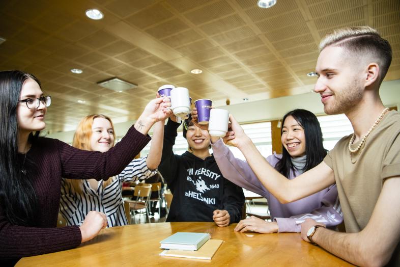 Students lifting cups of coffee and tea in the air at a table on the Viikki campus.