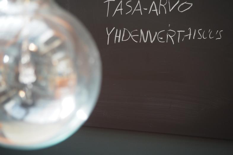 En lampa i förgrunden, en skrivtavla i bakgrunden med orden "likabehandling" och "jämställdhet" skrivna på finska.