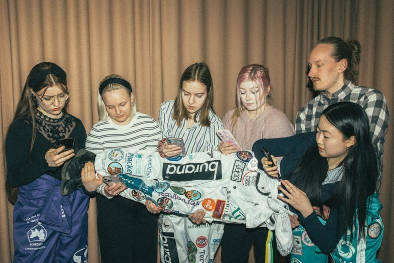 Five students browsing through their phones hold a sixth student, who is also holding a phone.