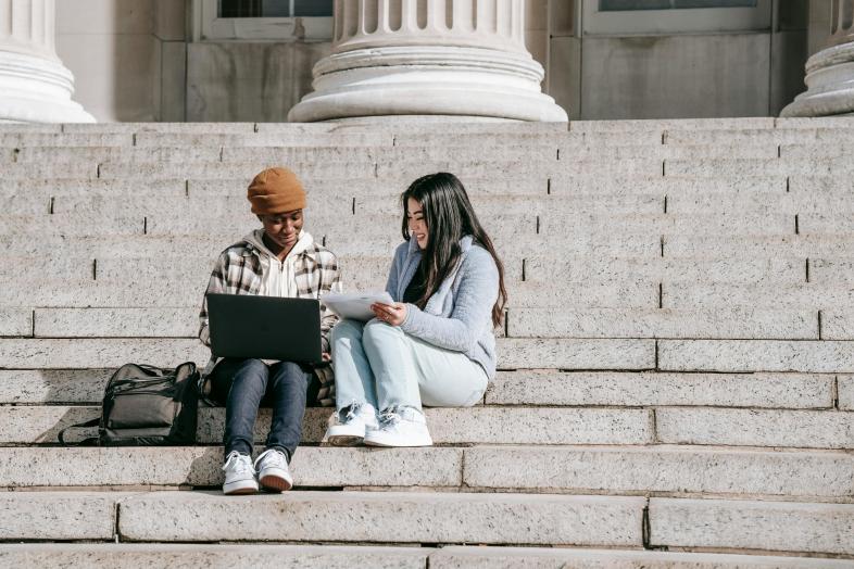 Two students sitting on a staircase with a laptop and some papers.