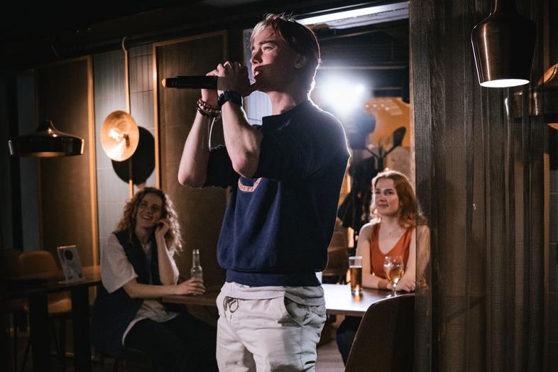 In the foreground a karaoke singer, in the background audience seated at a table.