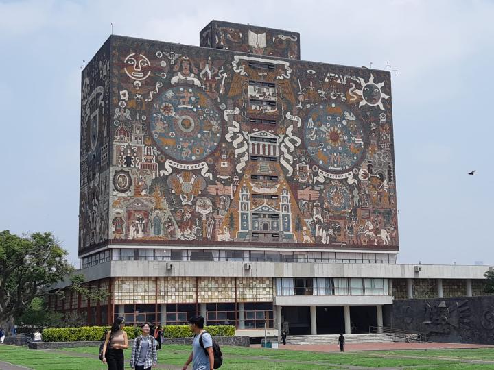 Tall building covered in colorful mosaics, grass, people walking.